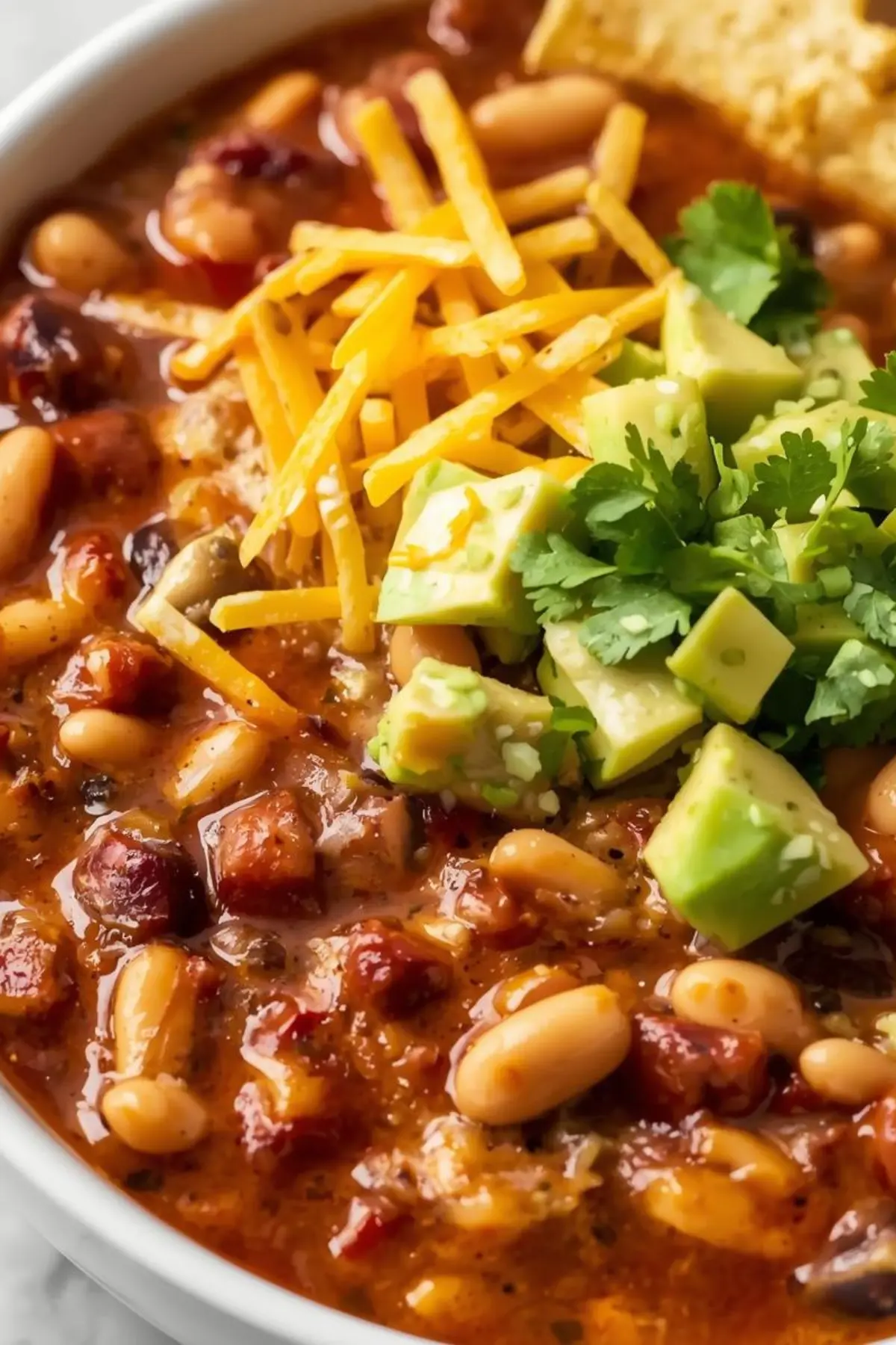 White bean chicken chili in a white bowl topped with cheese, avocado, cilantro and tortilla chips
